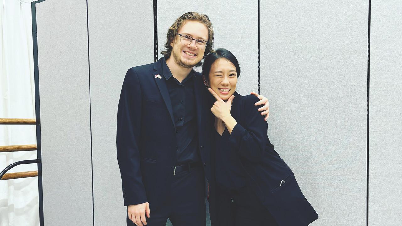 Two people, identified in the accompanying caption, stand side by side, smiling warmly. Both are dressed in black attire, and they stand against a light-colored background, exuding a sense of camaraderie and joy.